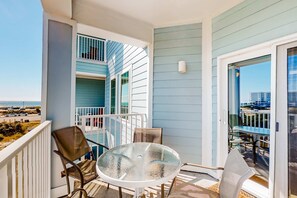 A balcony with a glass table and chairs, a view of the beach, and a building with blue siding. - Gulf view condo with pool & fitness center - balcony, open layout, and W/D (Orange Beach)