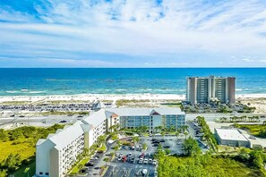 A coastal hotel with a parking lot, beachfront view, and a tall building in the background. - Gulf view condo with pool & fitness center - balcony, open layout, and W/D (Orange Beach)