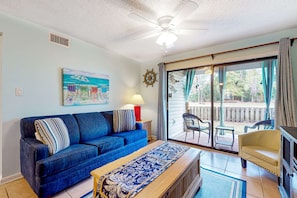 A living room with a blue sofa, a patterned rug, a ceiling fan, and a view through a sliding glass door to an outdoor patio. - Ground-floor condo with waterfront pool, private patio, AC, & W/D (Orange Beach)