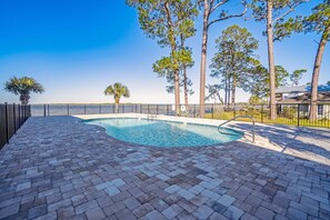 A swimming pool with a brick deck, surrounded by a fence and trees. - Ground-floor condo with waterfront pool, private patio, AC, & W/D (Orange Beach)