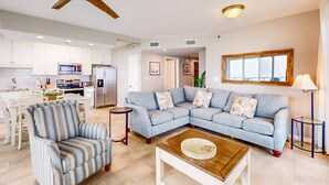 Una sala moderna con un sofá modular, un sillón a rayas, una mesa de centro, un ventilador de techo y una cocina al fondo.
