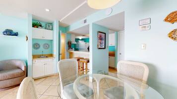 A living room with a sofa, a glass-top dining table with chairs, a kitchen area with a refrigerator, and a dining bar.