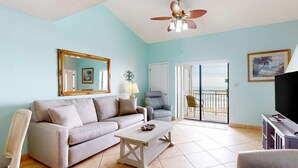 Un salon comprenant un canapé, une table basse, un meuble pour la télévision, un ventilateur de plafond, une porte donnant sur un balcon et un grand miroir.