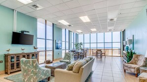 A spacious living area with a large window, a flat-screen TV, and a dining area with a view. - Gulf-front fourteenth-floor condo with pools, hot tub & great view (Orange Beach)