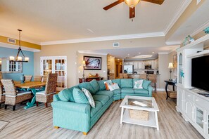 A spacious living room with a sectional sofa, a dining area with wicker chairs, and a kitchen in the background. - Coastal getaway with beach access, pool, hot tub, lazy river, & balcony (Orange Beach)