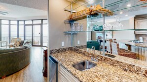 A modern kitchen with granite countertops, a stainless steel sink, and a glass display shelf. - Coastal getaway with beach access, pool, hot tub, lazy river, & balcony (Orange Beach)