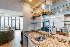 A modern kitchen with granite countertops, a stainless steel sink, and a glass display shelf. - Coastal getaway with beach access, pool, hot tub, lazy river, & balcony (Orange Beach)