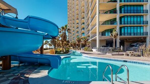 A swimming pool with a water slide, palm trees, and a multi-story building in the background. - Coastal getaway with beach access, pool, hot tub, lazy river, & balcony (Orange Beach)