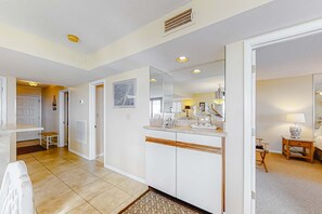 A modern kitchen with white cabinets, a countertop, and a large mirror. There is a door leading to another room and a small stool near the entrance. - Beachfront Views Beside Perdido Pass (Orange Beach)