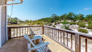 Eine Holzterrasse mit zwei Adirondack-Stühlen, einer Geländer und Blick auf eine sandige Landschaft mit Bäumen.