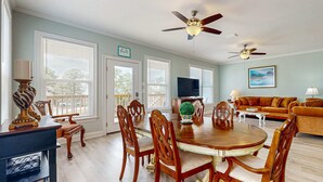 A dining area with a wooden table and chairs, a ceiling fan, and a television in the background. - Gulfview duplex with panoramic views, large balcony, and internet (Dauphin Island)