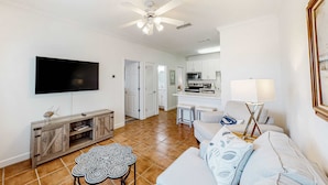 A living room with a TV, a ceiling fan, a dining area with a kitchen in the background, and a sofa with a lamp. - Newly renovated second-floor condo with shared pool, WiFi, W/D, & central AC (Dauphin Island)