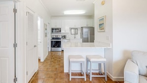 A kitchen with white cabinets, a central island, two bar stools, and a refrigerator. - Newly renovated second-floor condo with shared pool, WiFi, W/D, & central AC (Dauphin Island)