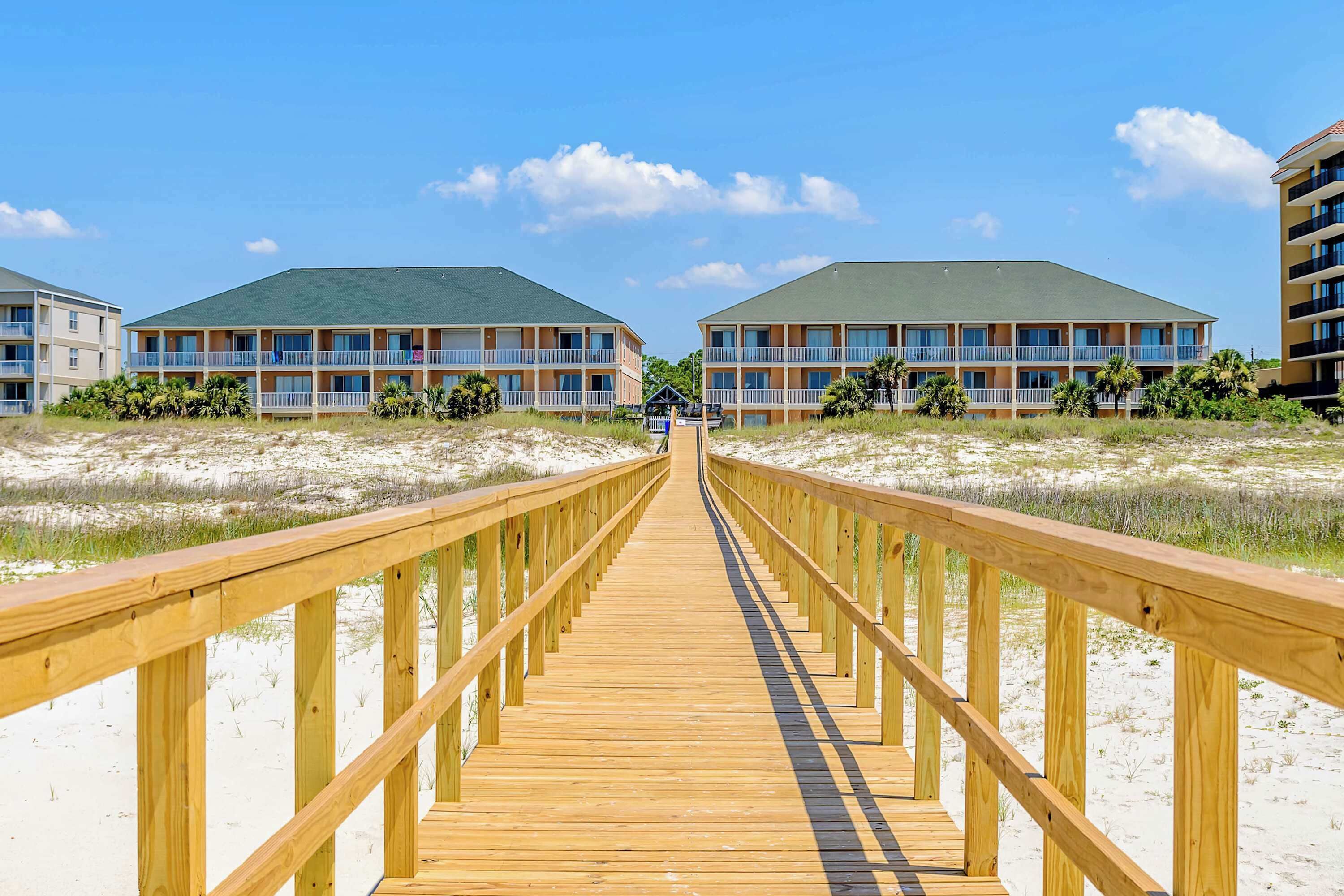 A wooden boardwalk leading to a beachfront hotel with multiple balconies and a clear sky.