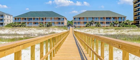 Ein Holzsteg führt zu einem Strandhotel mit mehreren Balkonen und blauem Himmel.
