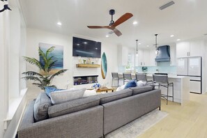 Een moderne woonkamer met een grijze bank, een eethoek met stoelen, een keuken met witte kasten en een plafondventilator.