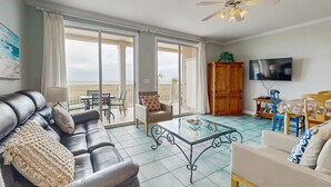 A living room with a sofa, armchair, coffee table, TV, and a balcony with outdoor furniture. - Gulf-Front Condo w/ Balcony, Shared Pools, and Beach Access (Dauphin Island)