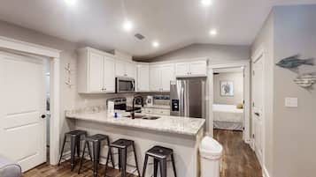 A compact kitchen with white cabinetry, a stainless steel refrigerator, a microwave, and a sink. There are three bar stools and a trash can.