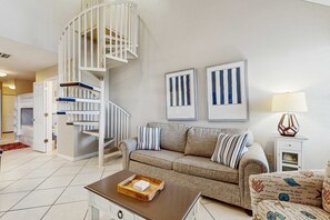 A living room with a beige sofa, a coffee table, a white spiral staircase, and framed artwork on the wall. - Cozy coastal condo with loft & shared pool and beach access (Orange Beach)