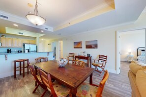 A dining area with a wooden table and chairs, a kitchen in the background, and a living room with a sofa. - Beachfront condo with pool, hot tub, W/D, sauna, balcony, and tennis court (Dauphin Island)