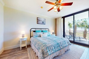 A bedroom with a bed, a ceiling fan, a nightstand, a lamp, a painting, and a balcony with chairs. - Beachfront condo with pool, hot tub, W/D, sauna, balcony, and tennis court (Dauphin Island)