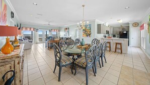 A spacious dining area with a glass-top table, surrounded by chairs with patterned upholstery, a chandelier, and a kitchen area with a refrigerator and sink. - Gulf front home with private dune walk over, internet, & washer & dryer (Dauphin Island)