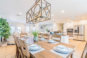 A spacious dining area with a wooden table set for four, surrounded by chairs. A decorative metal chandelier hangs above. - Bayfront beauty with a pool, grill, dock, reading room, board games, & AC (Dauphin Island)