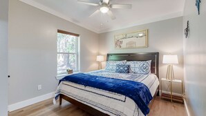 A bedroom with a bed, a ceiling fan, a window, a bedside lamp, and a wall-mounted artwork.