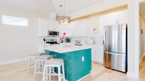 A modern kitchen with white cabinets, a stainless steel refrigerator, a central island with stools, and a hanging light fixture. - Beautiful beach house with Gulf views, covered deck, central AC, and free WiFi (Dauphin Island)