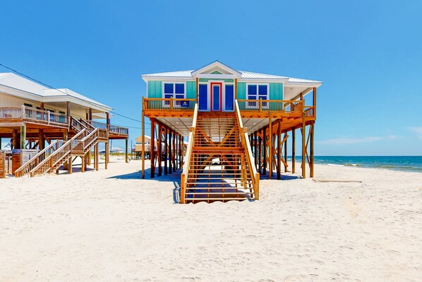 A beachfront house with a wooden staircase leading to a balcony, a pool, and a beach view.