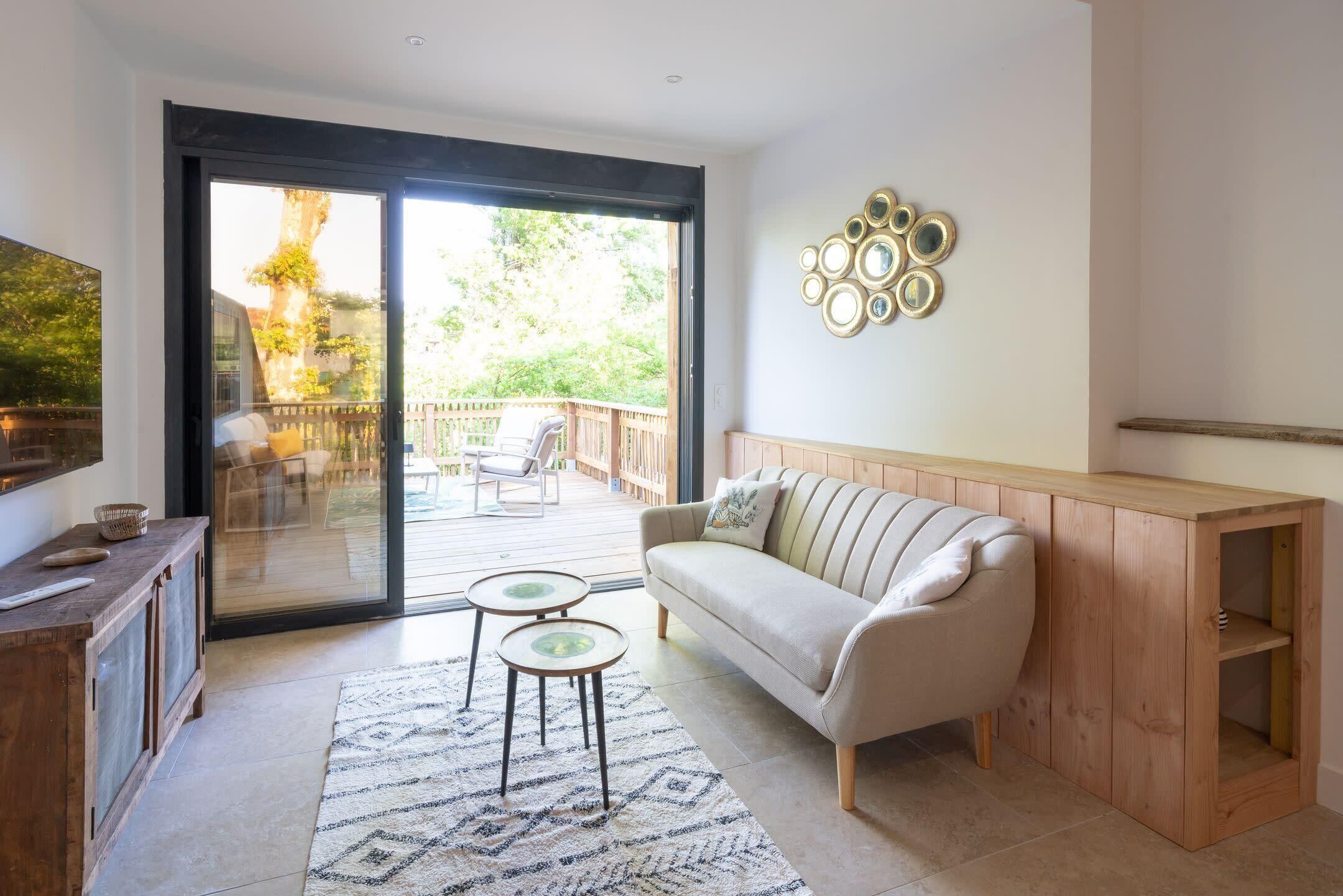 A living room with a sofa, a coffee table, and a TV on a wooden cabinet. There is a sliding glass door leading to an outdoor patio with a deck and chairs.