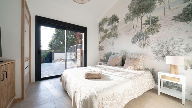 Une chambre avec un grand lit, une porte-fenêtre coulissante donnant sur un balcon, une fresque représentant des arbres et une table de chevet blanche.