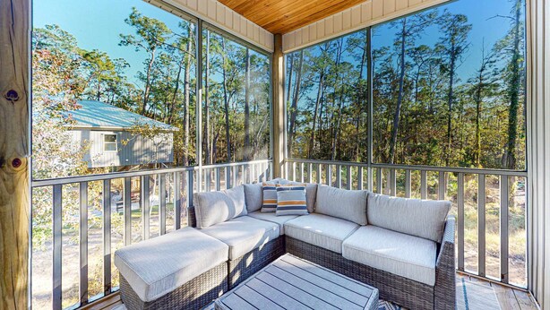 Eine überdachte Veranda mit einem Rattansofa, großen Fenstern und Blick auf Bäume sowie ein entferntes Gebäude.