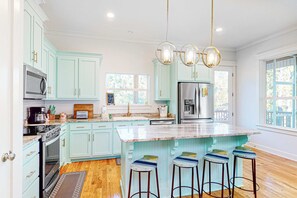Une cuisine avec des meubles de couleur bleu clair, un grand îlot central doté de tabourets, des appareils électroménagers en acier inoxydable et des luminaires suspendus.