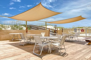 A covered outdoor seating area with tables and chairs, a wooden deck, and a view of the beach. - Gulf Front Getaway w/ Free WiFi, Central A/C, Balcony, Shared Pool, Beach Access (Orange Beach)