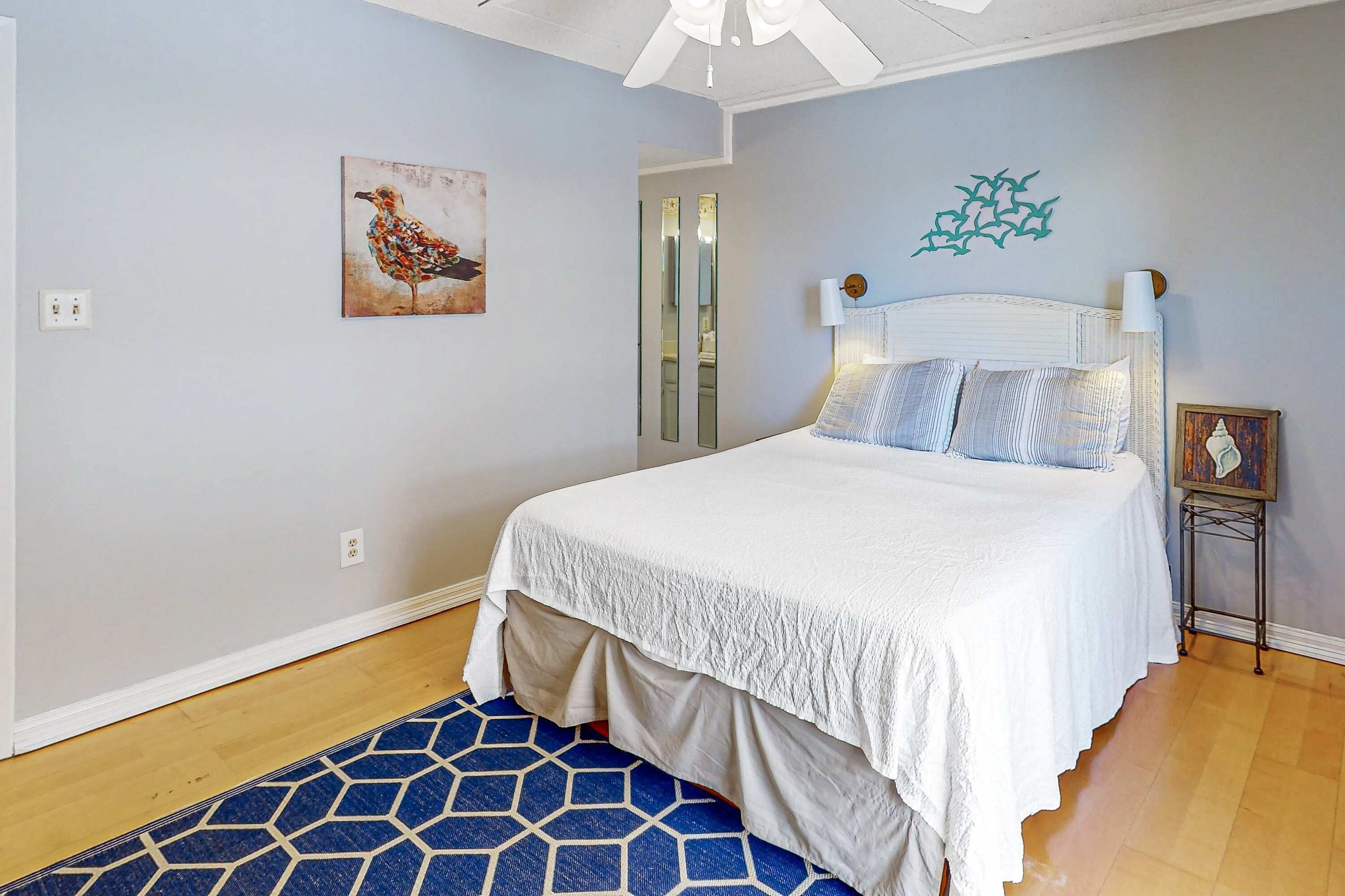 A bedroom with a bed, a blue patterned rug, a wall-mounted mirror, and a bird painting.