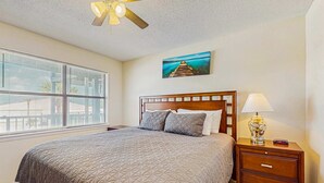 A bedroom with a bed, a nightstand, a lamp, a ceiling fan, a window with blinds, and a painting on the wall. - Condo with partial Gulf views, shared outdoor pool, sun deck & private beach access (Orange Beach)