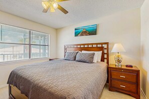A bedroom with a bed, a nightstand, a lamp, a ceiling fan, a window with blinds, and a painting on the wall. - Condo with partial Gulf views, shared outdoor pool, sun deck & private beach access (Orange Beach)