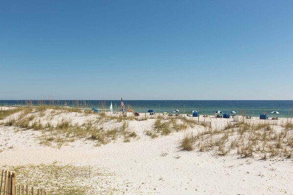 A beach with sand dunes, beach umbrellas, and a flag. - Condo with partial Gulf views, shared outdoor pool, sun deck & private beach access (Orange Beach)