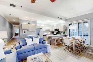 A spacious living room with a blue sofa, a dining area with a white table and chairs, and a kitchen with white cabinets and a large window. - Ground floor, Gulf front condo with pools, hot tubs, tennis courts, and gym (Orange Beach)