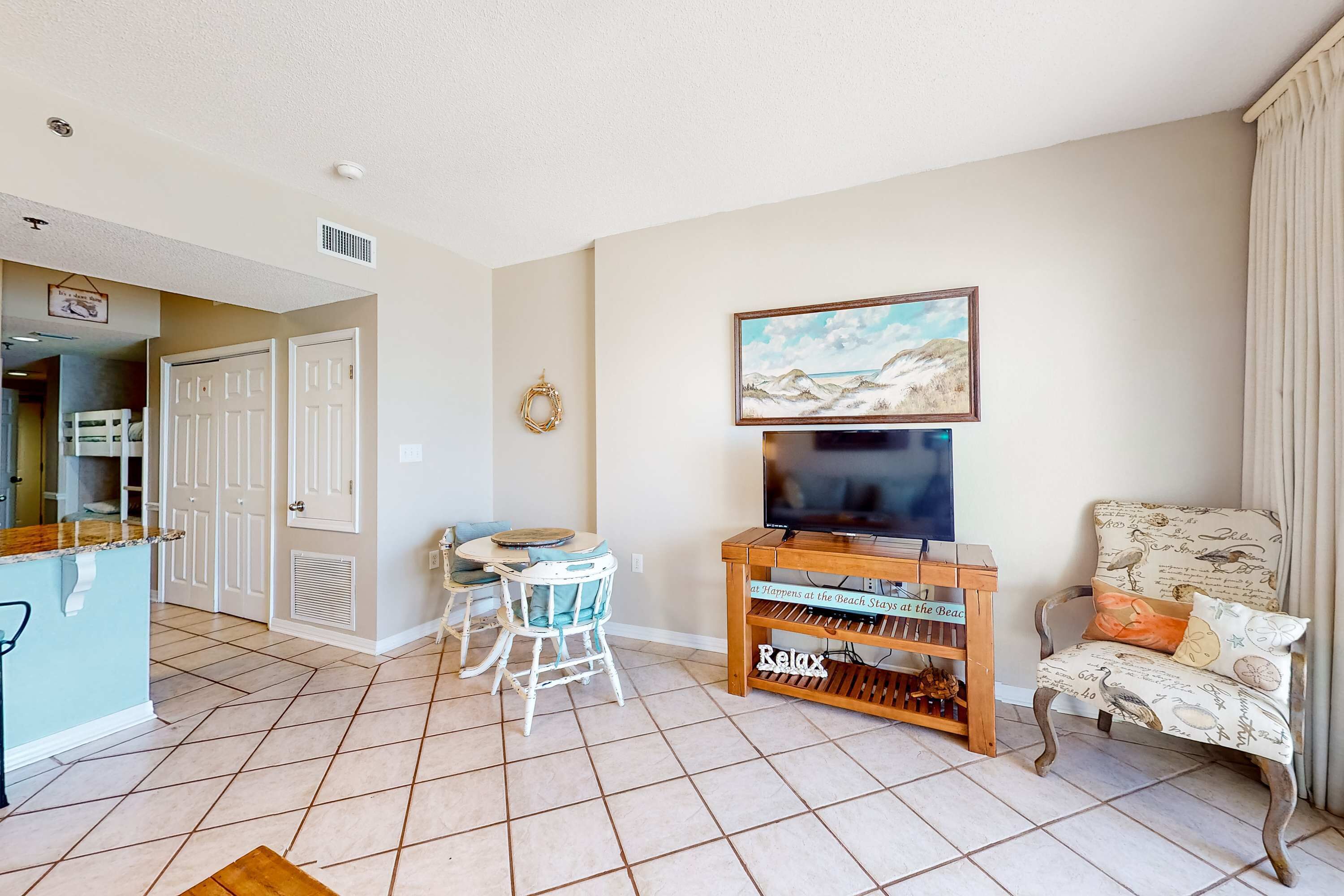 A living room with a TV, a dining area with chairs, a kitchen area, and a door leading to another room.
