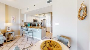 A modern kitchen with white cabinets, a central island, and a dining area with a table and chairs. - Coastal condo w/ shared, outdoor pool, indoor pool w/ hot tub, & relaxing sauna (Orange Beach)