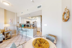 A modern kitchen with white cabinets, a central island, and a dining area with a table and chairs. - Coastal condo w/ shared, outdoor pool, indoor pool w/ hot tub, & relaxing sauna (Orange Beach)
