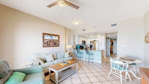 A living room with a sofa, coffee table, and a dining area with chairs and a table. There is a kitchen in the background with cabinets and appliances. - Coastal condo w/ shared, outdoor pool, indoor pool w/ hot tub, & relaxing sauna (Orange Beach)