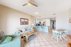 A living room with a sofa, coffee table, and a dining area with chairs and a table. There is a kitchen in the background with cabinets and appliances. - Coastal condo w/ shared, outdoor pool, indoor pool w/ hot tub, & relaxing sauna (Orange Beach)