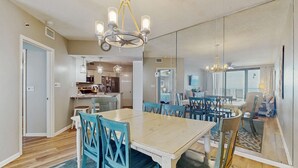 Un comedor con una mesa grande y sillas, una araña y una cocina al fondo.