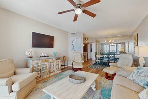 A living room with a ceiling fan, a flat-screen TV, a dining area with a table and chairs, and a kitchen in the background. - Contemporary Gulf front condo w/ shared pools, hot tub, fitness center, beach (Orange Beach)