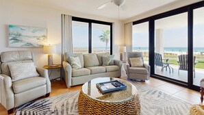 A spacious living room with a large window offering a view of the ocean, a ceiling fan, and a comfortable seating area with a coffee table. - Stunning Gulf front condo w/ a spacious layout, shared pools, & fitness room (Orange Beach)