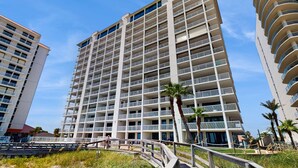 A multi-story hotel with balconies, a pedestrian bridge, and palm trees. - Stunning Gulf front condo w/ a spacious layout, shared pools, & fitness room (Orange Beach)