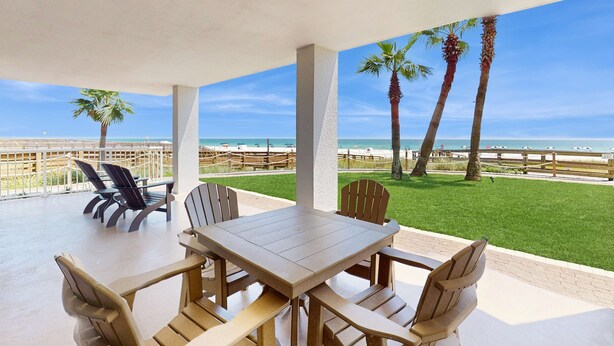 A covered outdoor seating area with a table and chairs, overlooking a beach with palm trees and a fence. - Stunning Gulf front condo w/ a spacious layout, shared pools, & fitness room (Orange Beach)